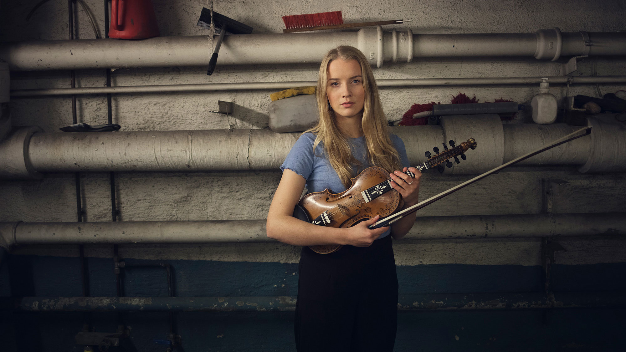 Helga Myhr står med fela foran industriell vegg