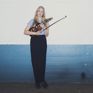 Helga Myhr stand in front of concrete wall with her Hardanger fiddle.