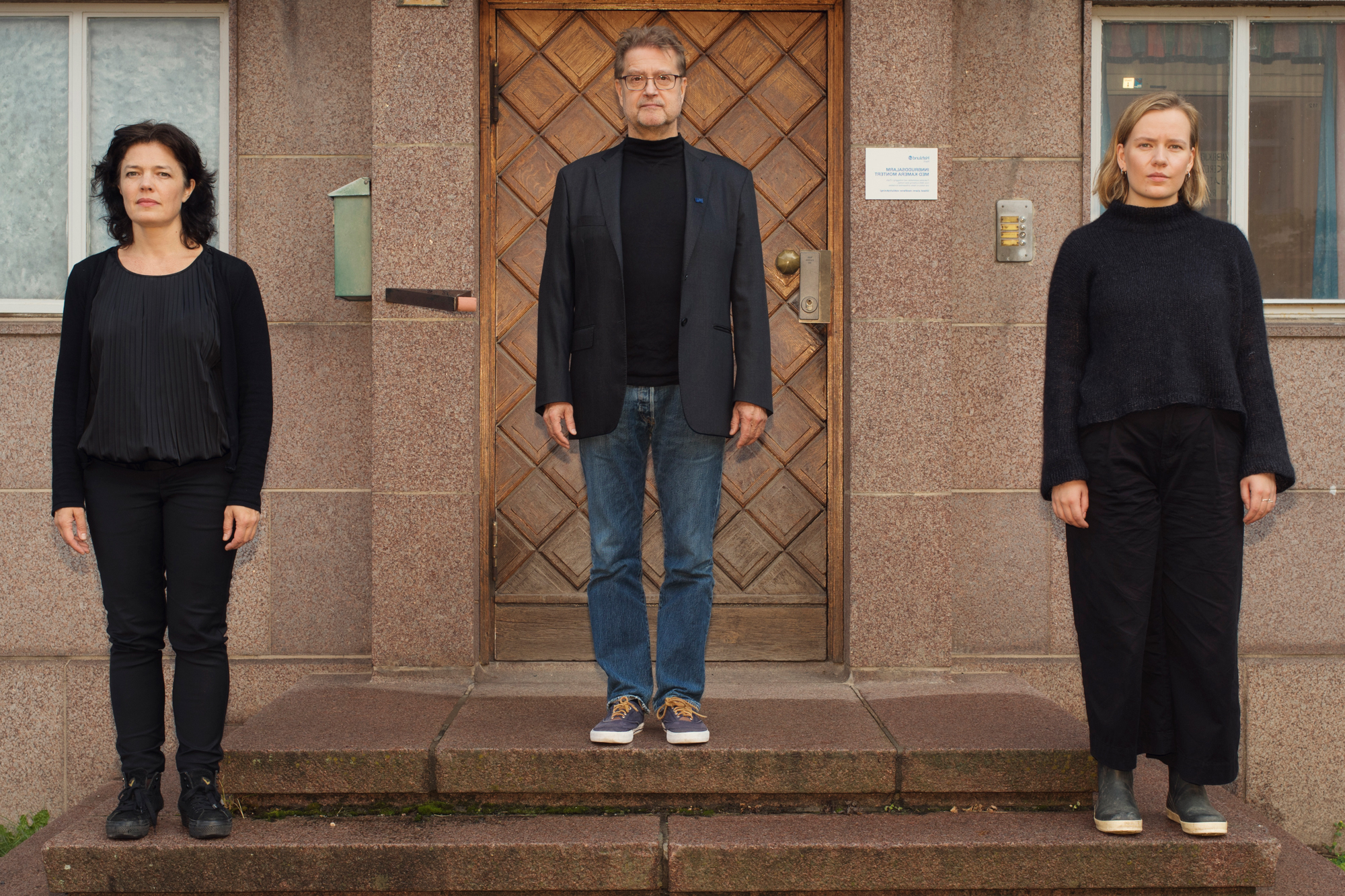 En mann omkranset av to kvinner står symmetrisk stilt opp utenfor en blokk i sommerlyset