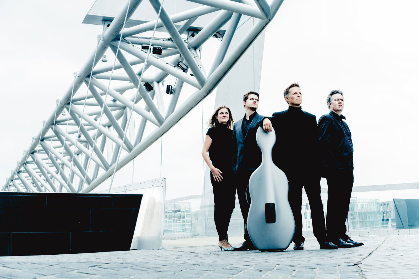 En dame og tre menn står på en bro i Bjørvika i Oslo og ser mot horisonten. En av dem lener seg mot en cello.