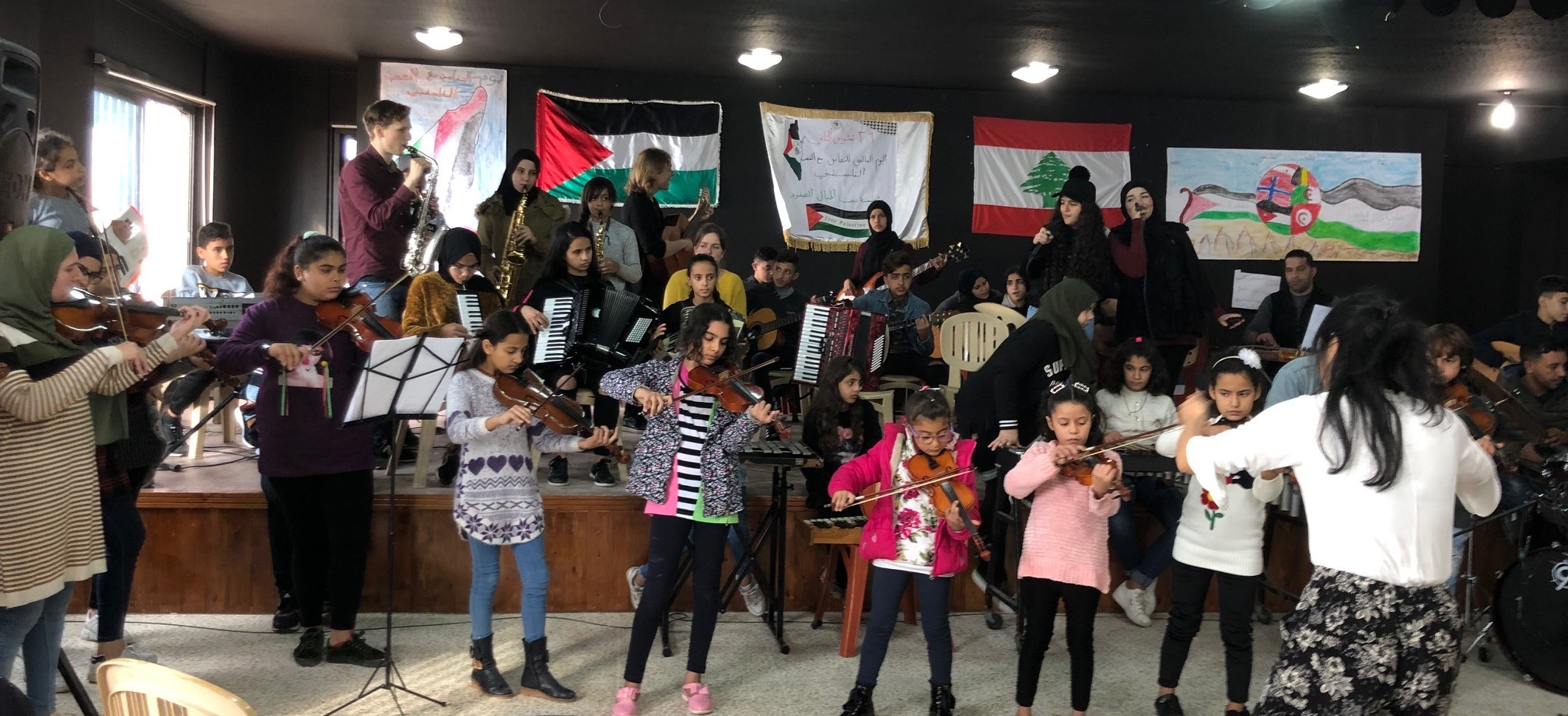 Barneorkester. Små barn står foran på rekke og spiller fiolin. Bak spiller større barn flere intrumenter på en scene. Flagg og bannere. Dirigent med ryggen til.