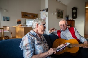 An elderly man and woman are sitting in a sofa. The woman is reading something from an iPad and the man are sitting with a guitar in his lap, smiling.
