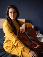 Cellist María Alejandra Conde Campos sitter på sengen med celloen