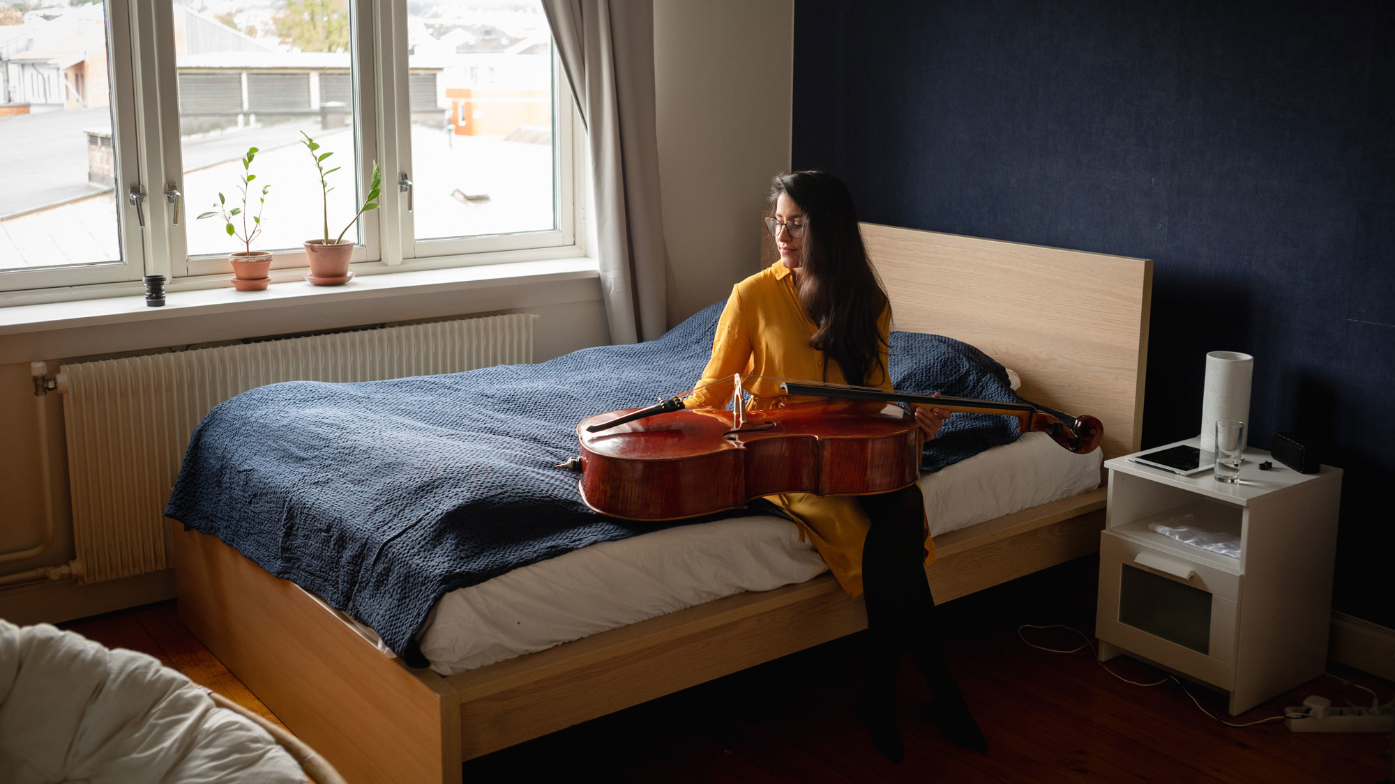 Cellist María Alejandra Conde Campos sitter på sengen med celloen på fange og ser vekk