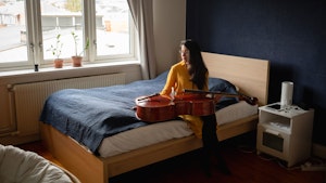 Cellist María Alejandra Conde Campos sitter på sengen med celloen på fange og ser vekk