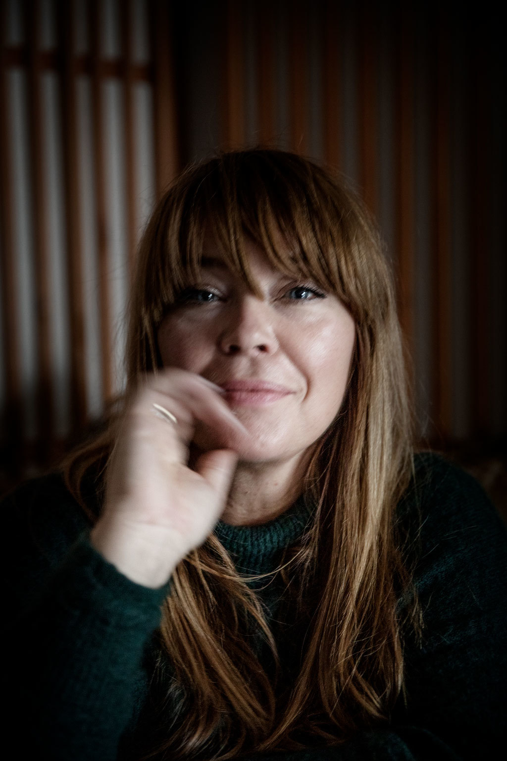 Solveig ser mot kameraet og fører en hånd mot ansiktet. Hun har tung pannelugg og smiler lett.