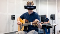 Johannes Lunde Hatfield sitter og spiller gitar med VR-briller på. Pål Hieronimus Aamodt sitter i bakgrunnen bak PC'en