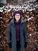 Astrid Kvalbein i helfigur foran en snødekt hekk med oransje vinterblader. Astrid har lilla alpelue og mørk grå boblejakke og smiler mot kamera.
