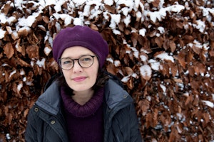 Astrid in front of snow-covered hedge smiling towards the camera. She is wearing a beret, round spectacles and a winter jacket.