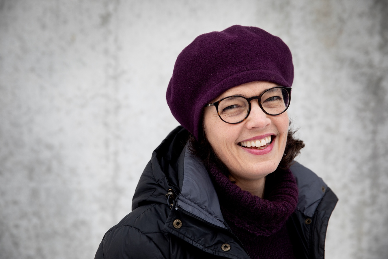 Astrid står mot en murvegg, ikledd lue og boblejakke, og smiler bredt mot kameraet.