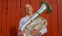 Øystein Baadsvik står foran en rød vegg og smiler, men tubaen i hendene.