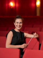 Nina Haug sitter i rød konsertsal med dirigentpinnen i hånda og smiler.