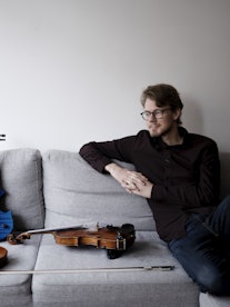 Jørgen Mathisen i grå sofa med tre fioliner ved siden av seg