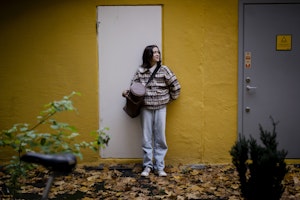 Kristina Fransson foran en hvit dør på gul vegg, med trompetveske i hånda