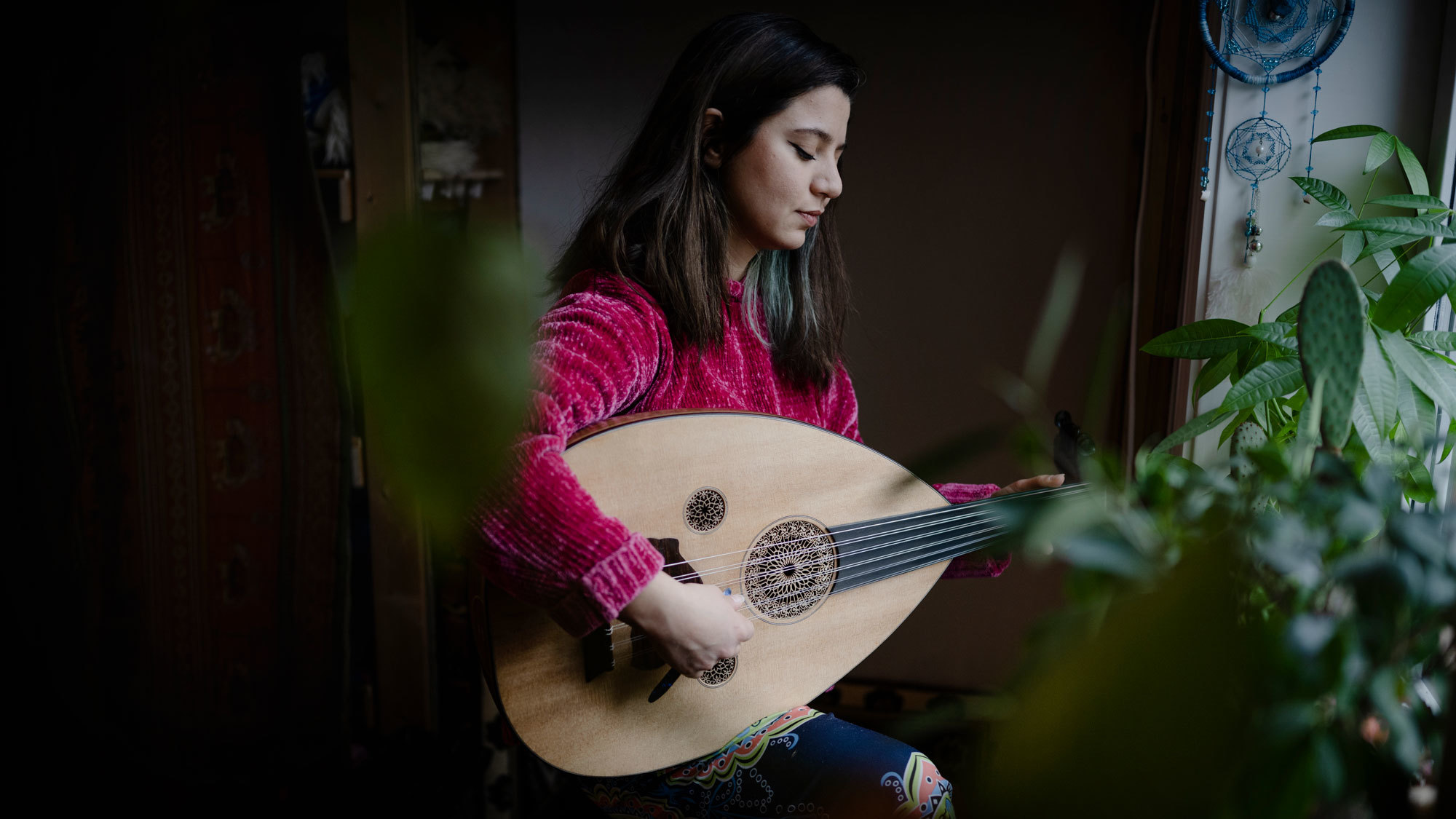 Nawar Alnaddaf i rosa genser som spiller oud mellom grønne planter på rommet sitt.