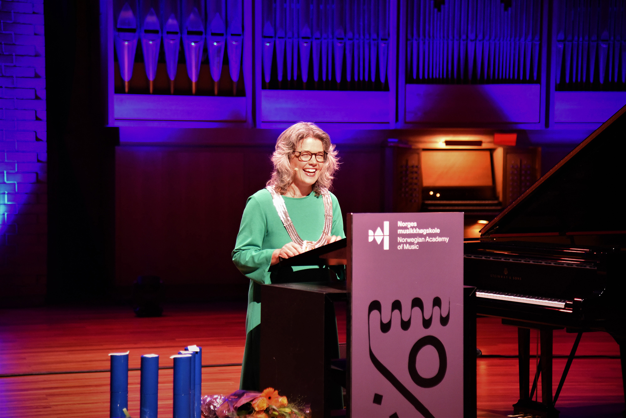 Astrid Kvalbein på talerstolen i Lindemansalen pyntet til fest. Grønn kjole og rektorkjede. Smilende.