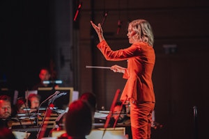 Hannah Howard i rød dress står foran orkesteret med dirigentstav i hånda. Musikere i bakgrunnen.