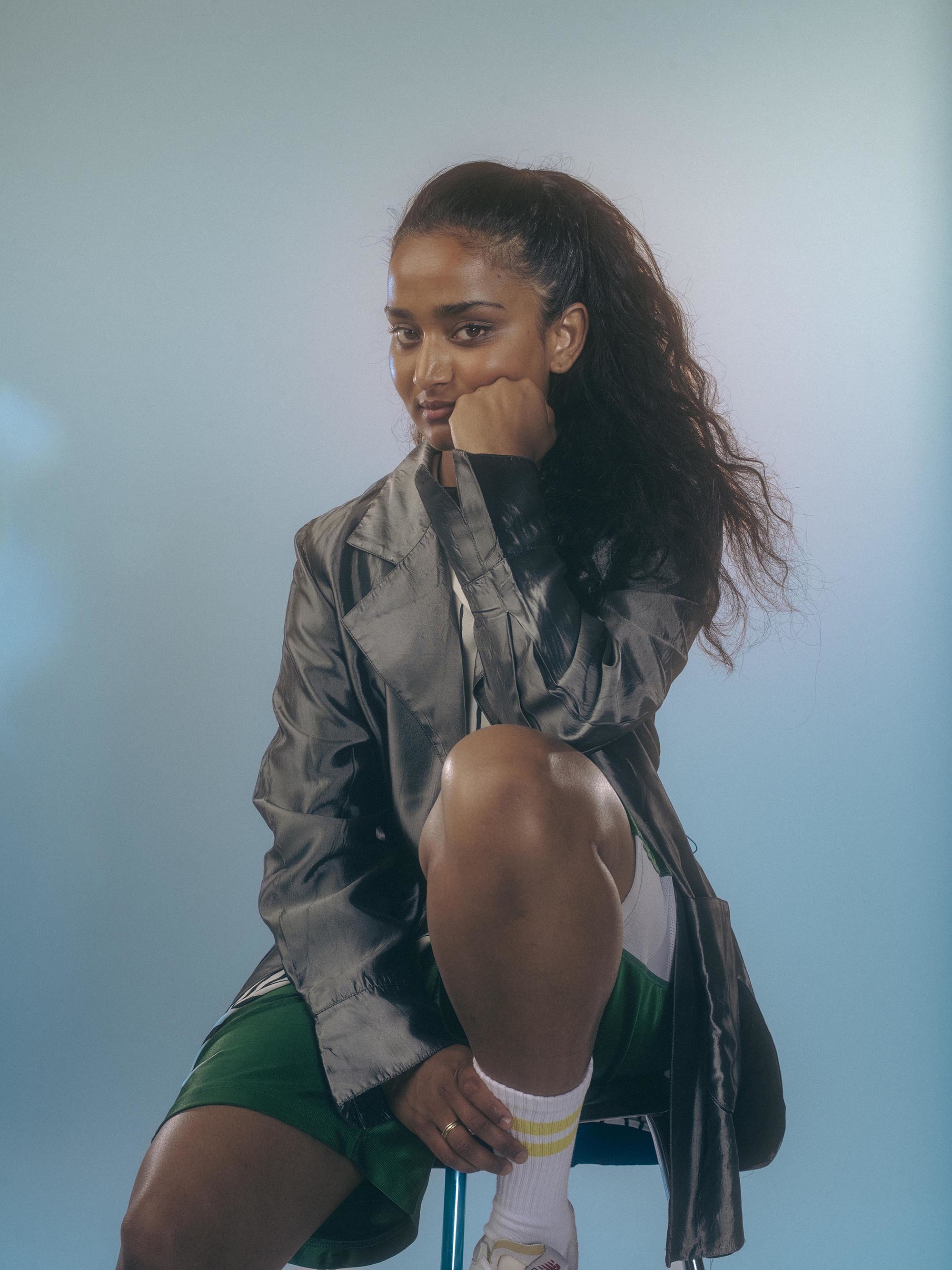 Mira Thiruchelvam i grå dressjakke og grønn kortbukse sitter på en krakk med en fot opp og den andre i gulvet. Blå bakgrunn.