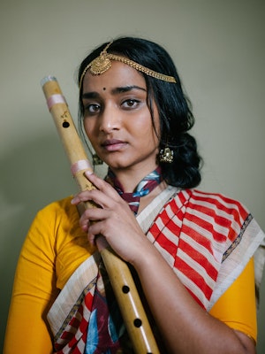 Mira Thiruchelvam i folkedrakt holder en fløyte opp på skrå. Halvfigur forfra. Alvorlig uttrykk.