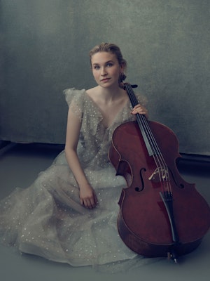 Helfigurbilde av Sophie Kauer i duse farger, der hun sitter på gulvet i en gammeldags kjole og celloen foran seg.