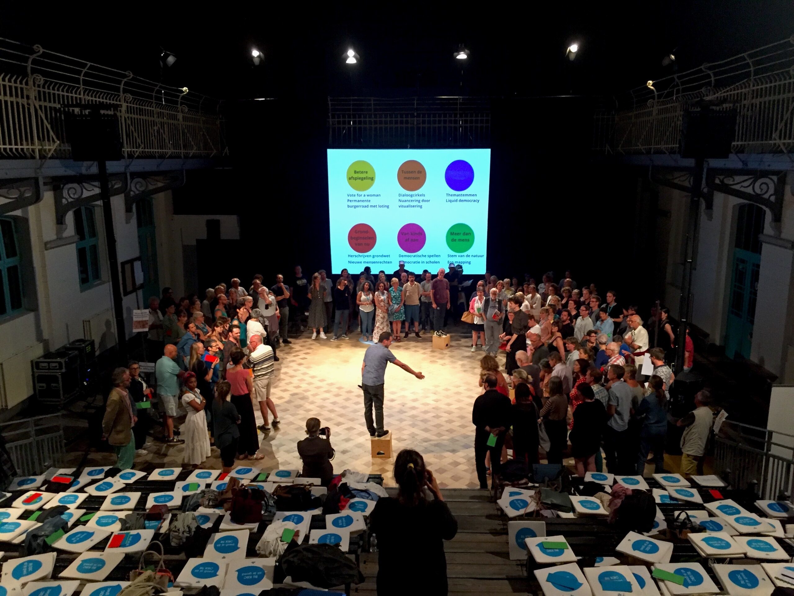 Dette pressebildet er fra en forestilling "In Search of Democracy 3.0.", og viser et scenegulv, et mørkt rom/black box, med masse folk som står i ring rundt en mann oppå en kasse, hvor han peker og taler.