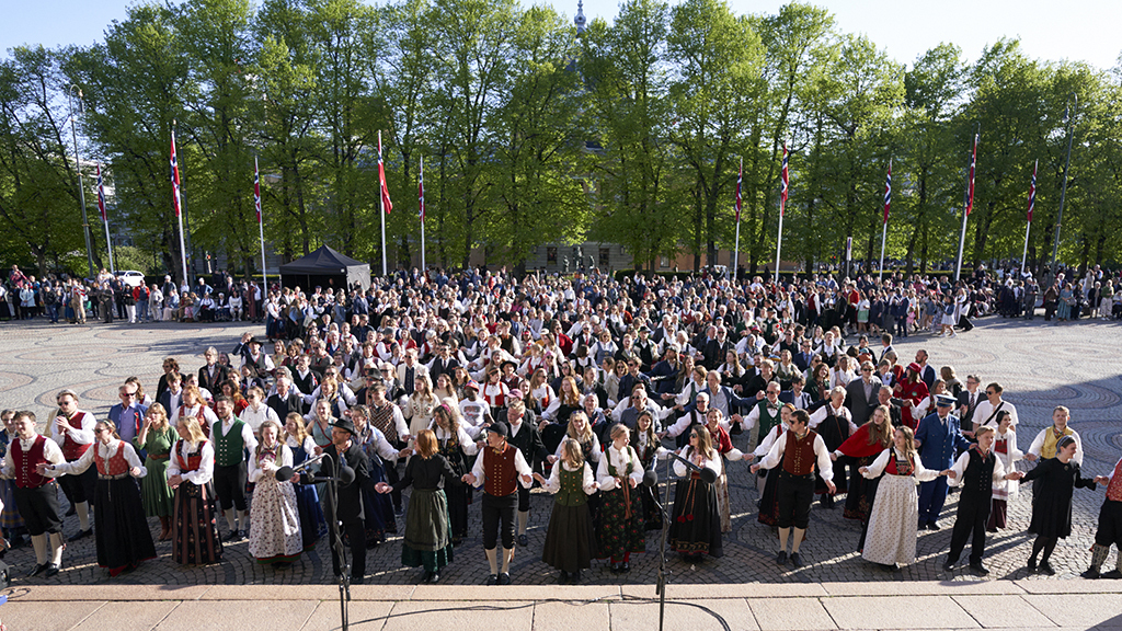 Mange mennesker i bunad holder hender foran trapp på Universitetets aula