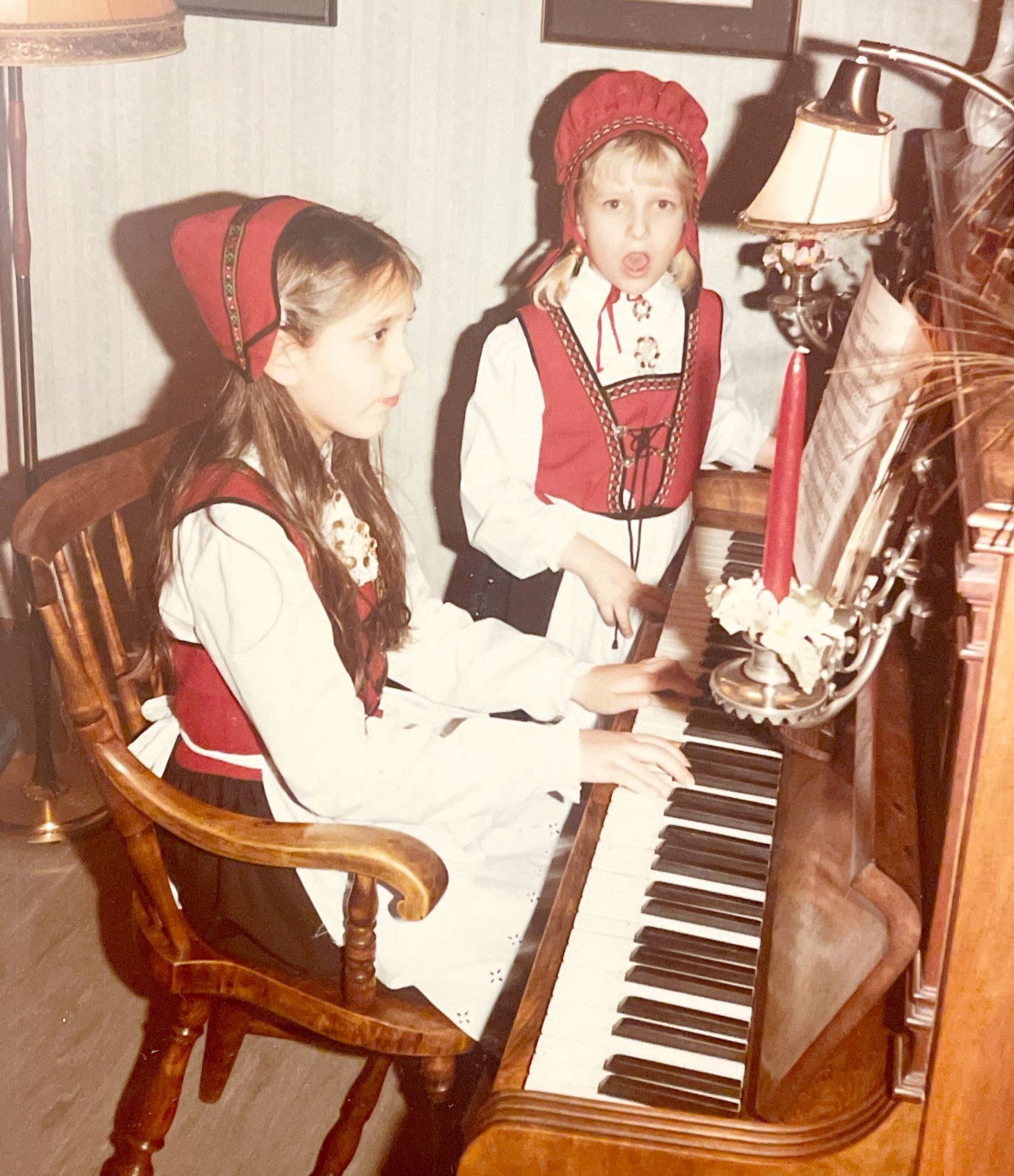 To jenter i bunad ved et brunt piano. En spiller og en synger.