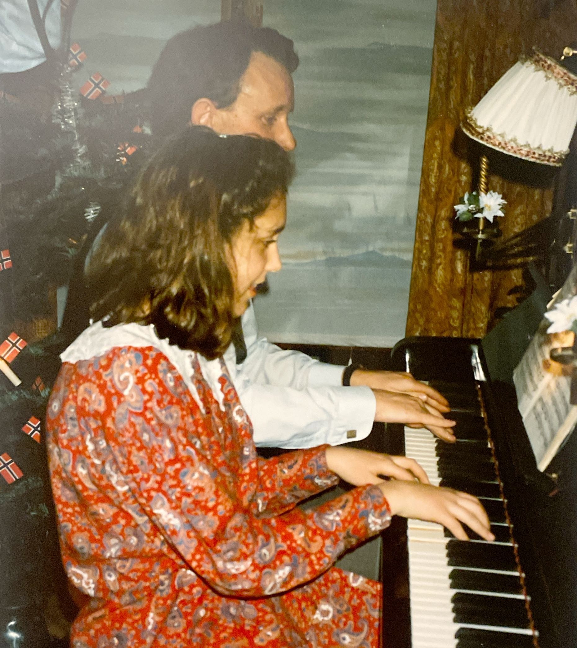 En mann og en ung kvinne spiller piano sammen, med et juletre i bakgrunnen.