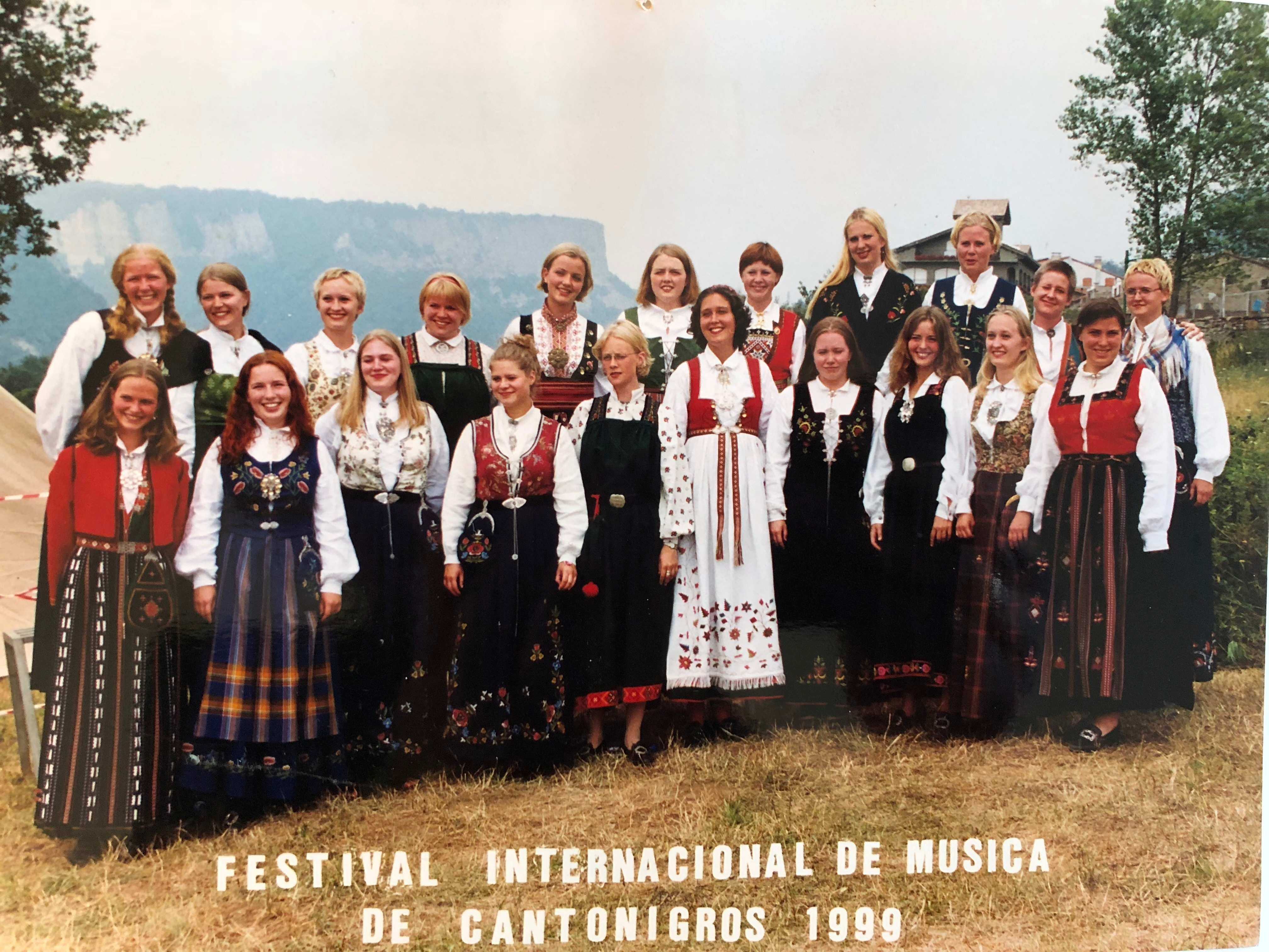 Kvinnekor i bunad står oppstilt på en plen med fjell og himmel i bakgrunnen.