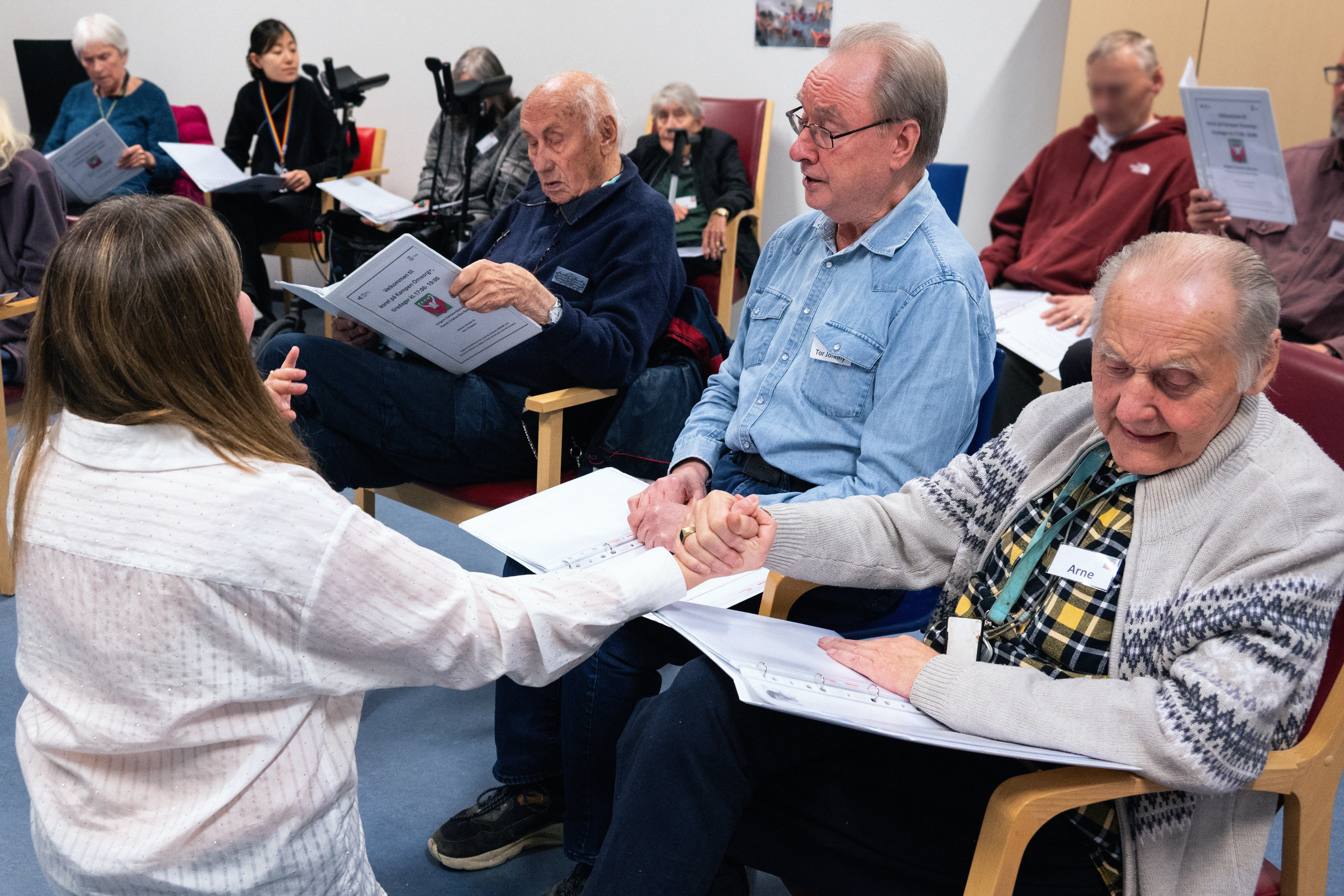 Korøvelse med eldre mennesker. I fokus er en ung kvinnelig dirigent som holder en eldre mann i hånden.
