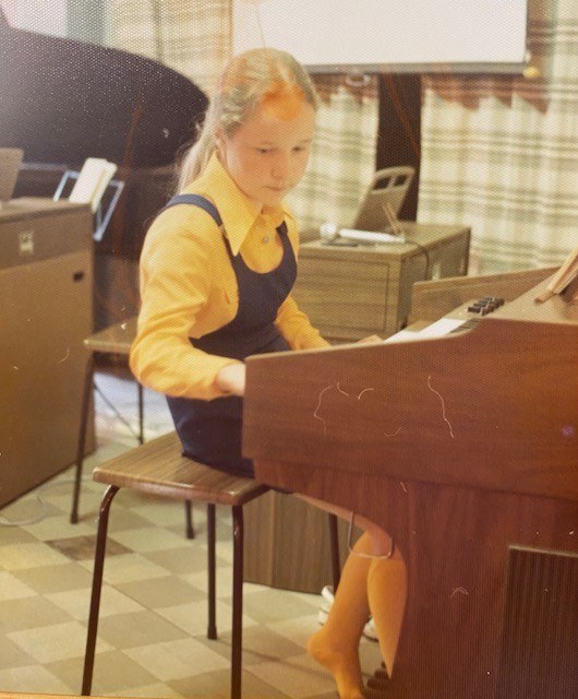 Jente sitter ved et el-orgel, med møbler og klær fra 70-tallet