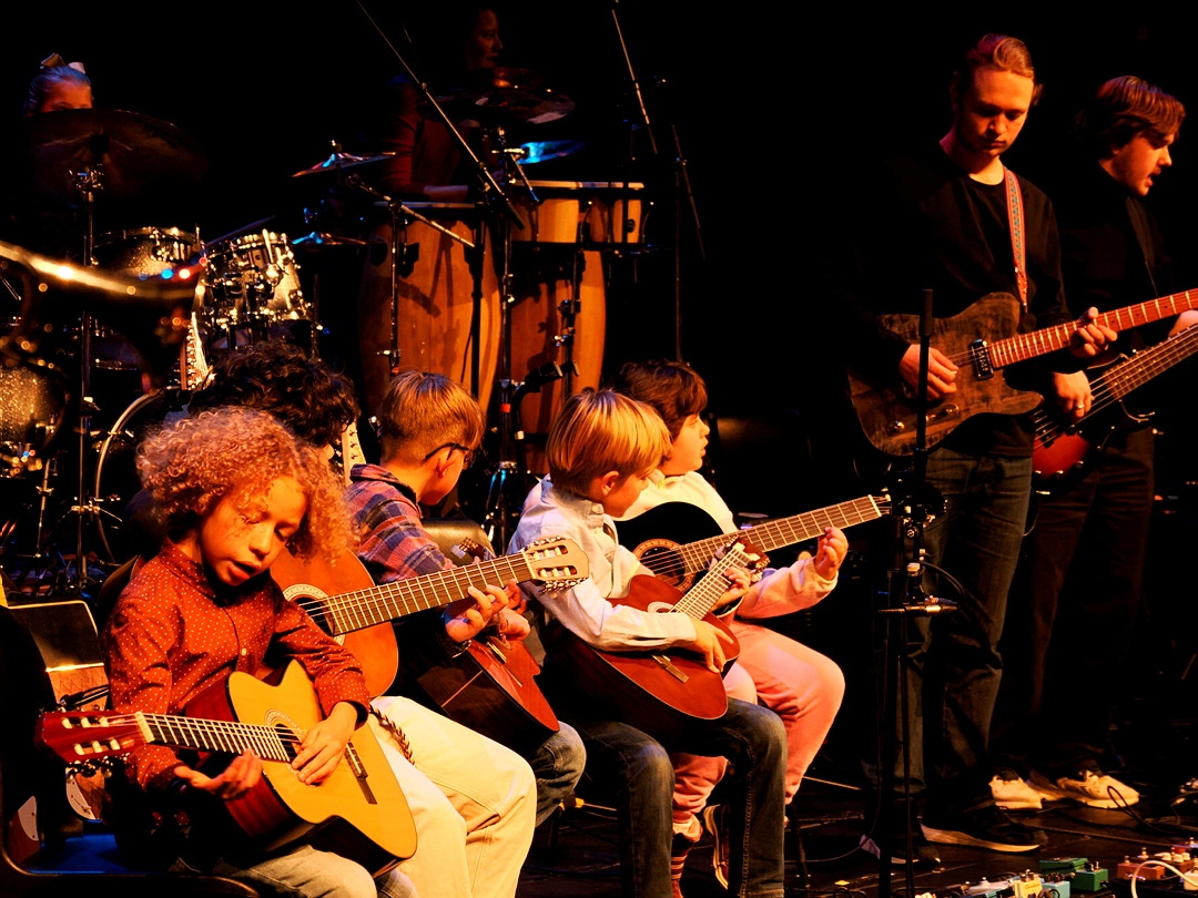 Små barn spiller gitar på en scene, mens voksne musikere spiller sammen med dem.
