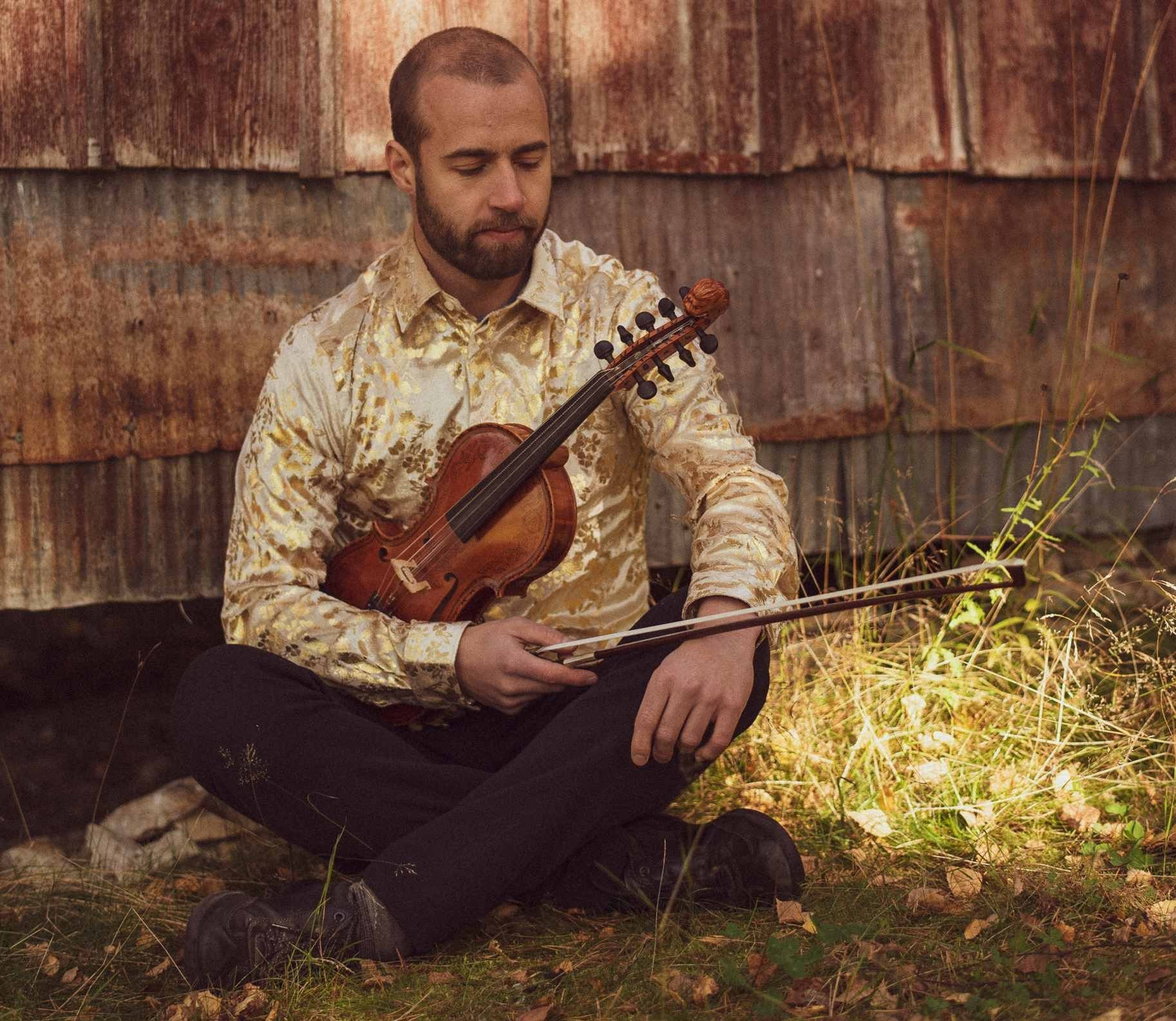 Sivert Holmen sitter på gresset foran en låve, med fela i fanget