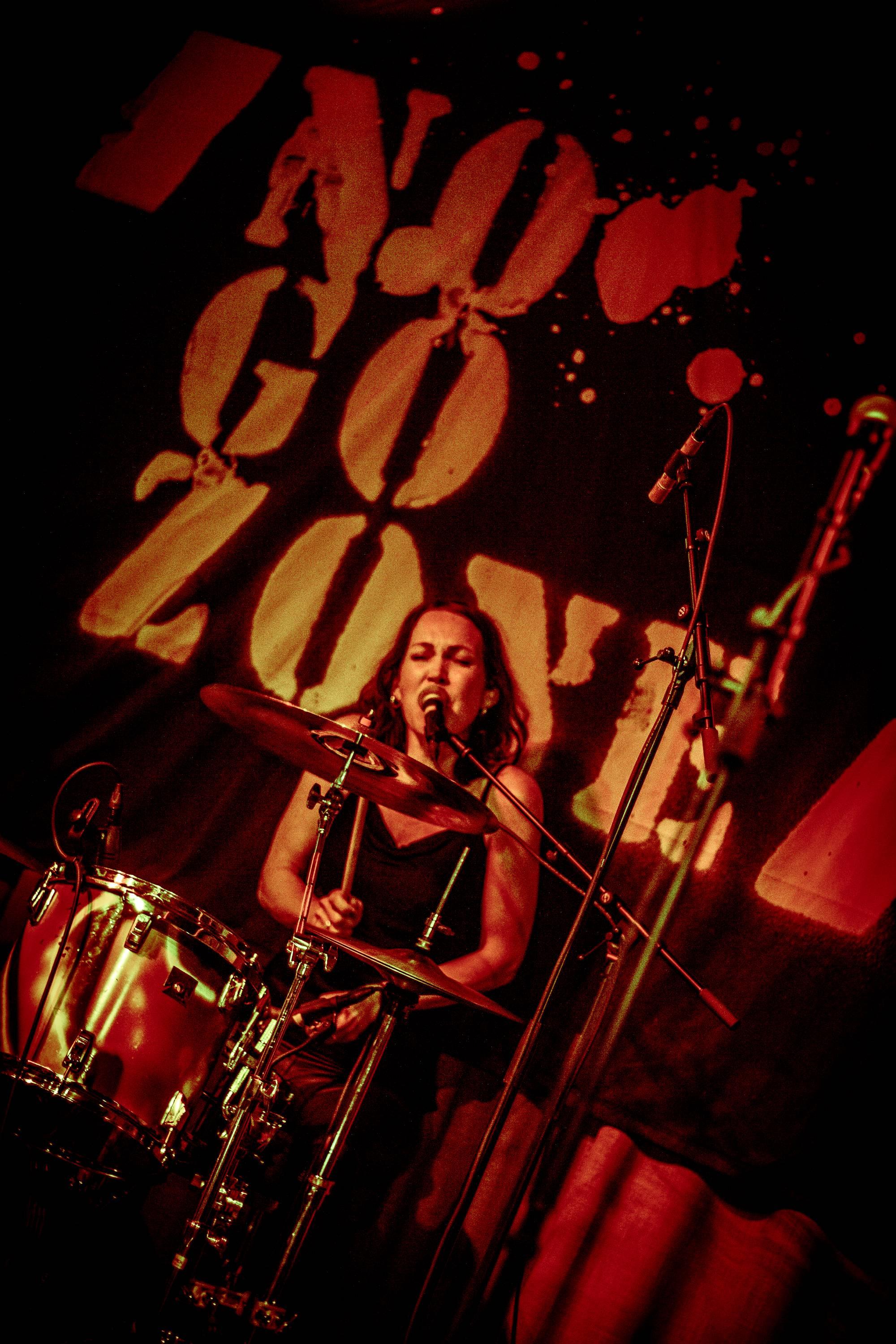 Kvinne, Lise Lotte Berg Ågedal, spiller trommesett i band, i et mørkt lokale med teksten "No Go Zone" på veggen bak