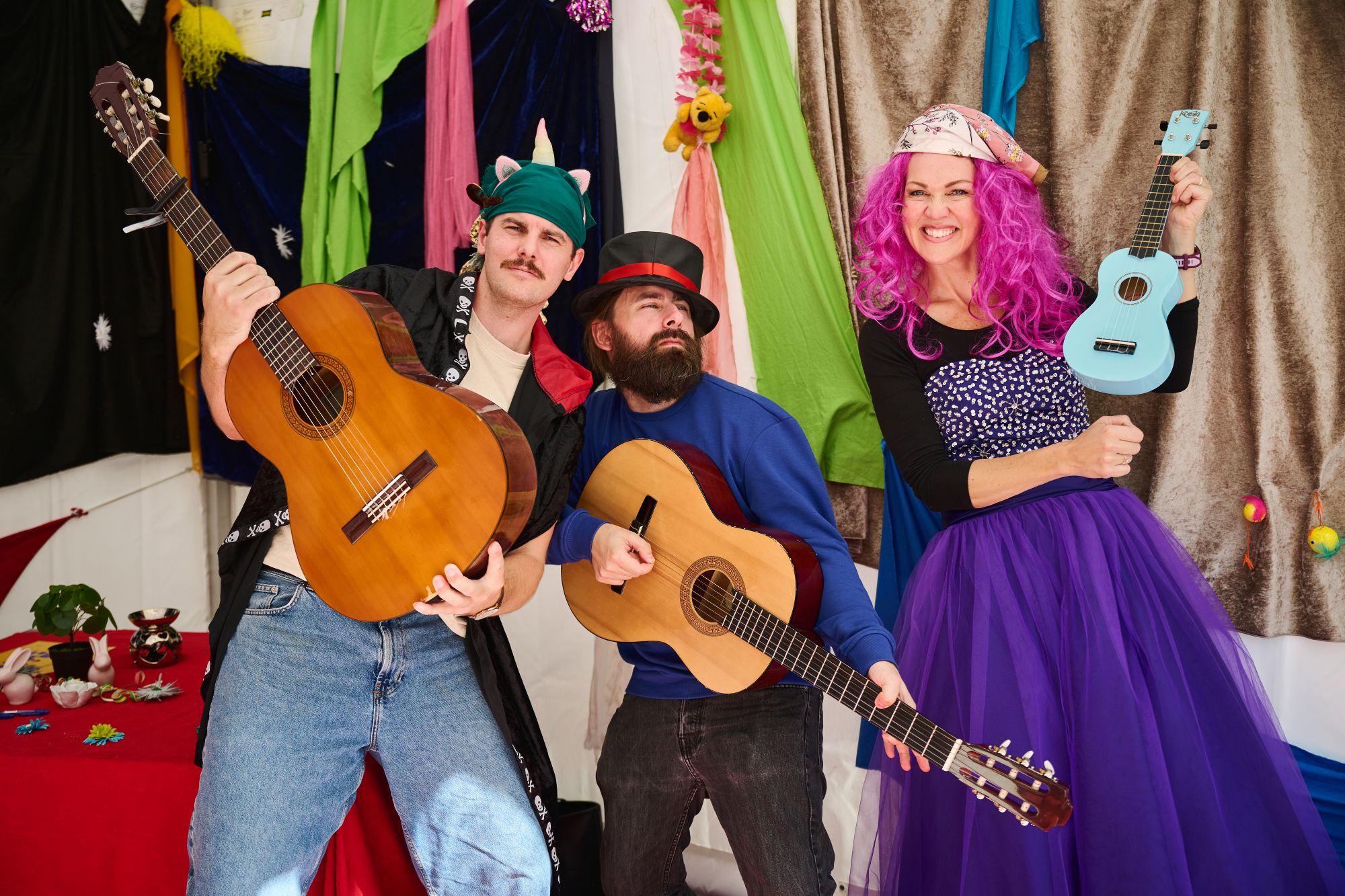 Tre personer med gitarer poserer som et band, alle har gitarer og er iført fargerike klær og hatter. En kvinne, Lise Lotte Berg Ågedalstår til høyre med rosa parykk.
