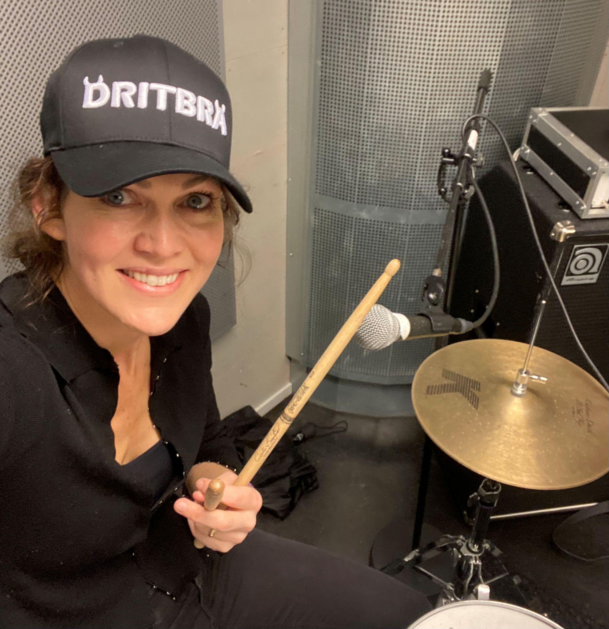 Kvinne, Lise Lotte Berg Ågedal, spiller trommesett i et lyst lokale iført en caps med teksten "Dritbra" på