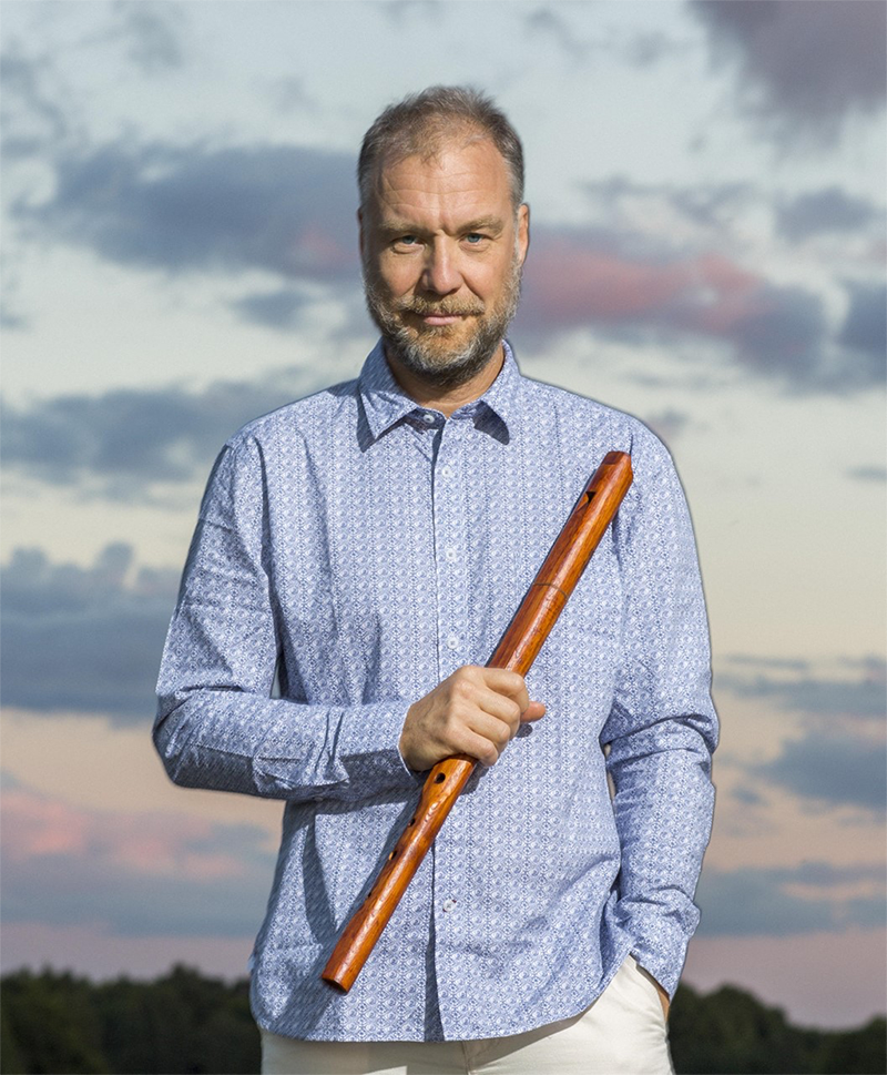 Mann med lyseblå skjorte holder en fløyte. Himmel i bakgrunnen.