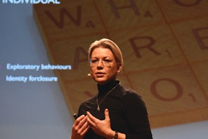 Kvinne med blondt hår og mikrofon står foran skjerm med hendene løftet