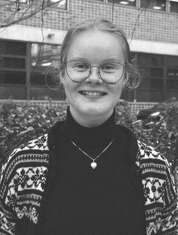 Svart/hvitt bilde av kvinne med lyst hår og briller i strikketcardigan og mørk genser foran bygning.
