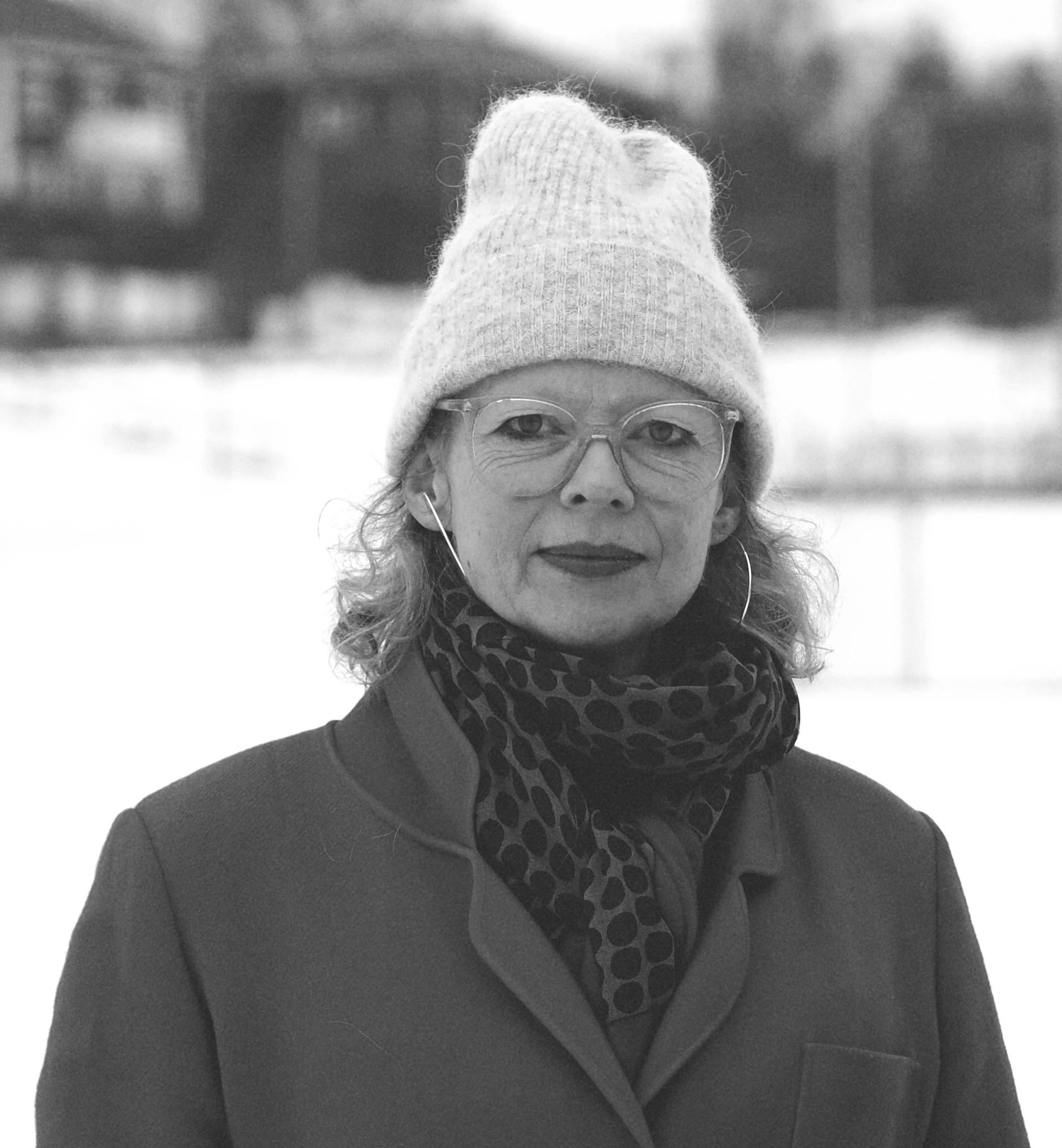 Sorthvitt bilde av kvinne med lue, briller, skjerf og jakke. Snø i bakgrunnen.