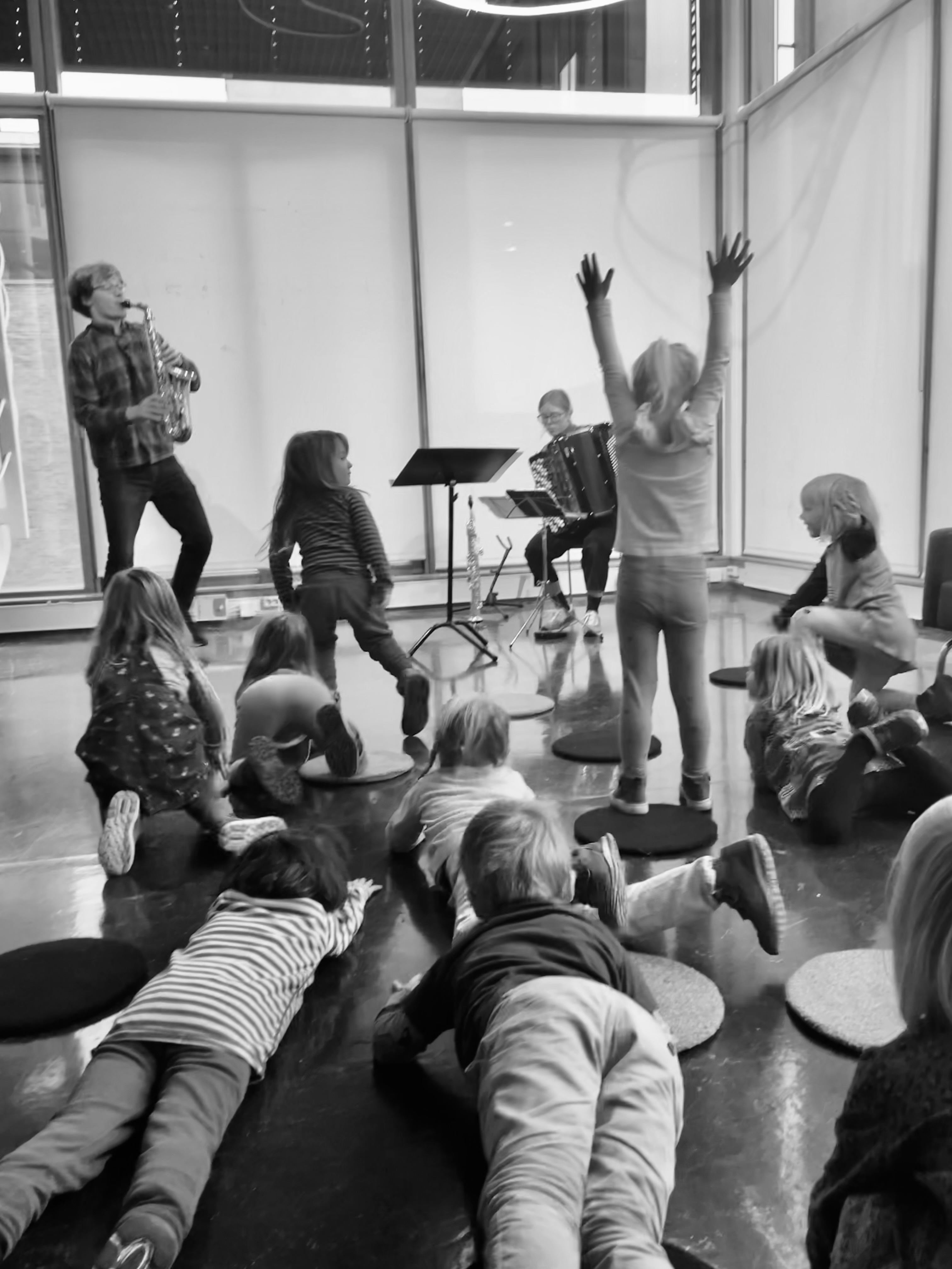 saksofonist og akkordeonist spiller mens barn strekker seg i været og på gulvet. Sort hvitt.