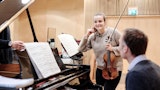 En student står ved siden av et flygel og smiler. Hun har en fiolin i hånda og ved pianoet sitter en akkopagnatør.