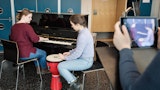 En student sitter og spiller piano, en spiller på en tromme og en tredje filmer med et nettbrett.