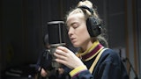 En student står å synger konsentrert inn i en mikrofon. Hun har store hodetelefoner på seg.