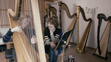 En studenter sitter smilende og spiller på en stor harpe. Læreren sitter rett ved siden av og smiler.