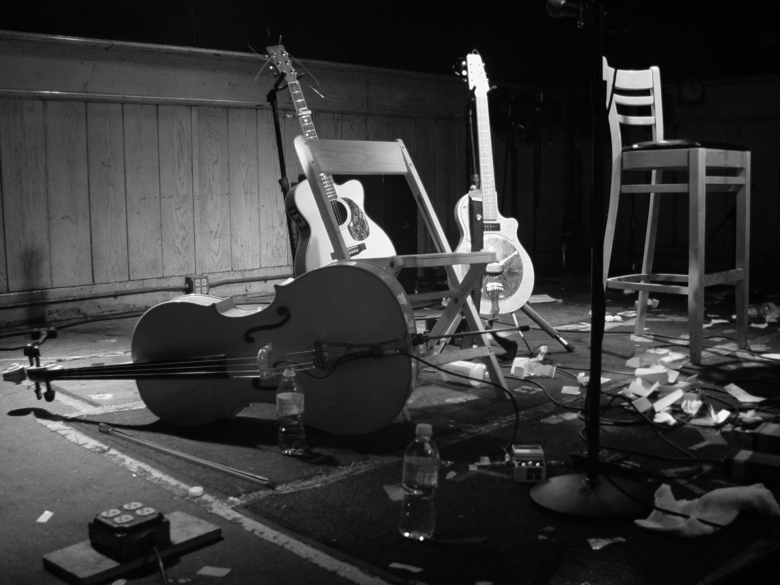 An electric and an acoustic guitar placed on guitar stands on a stage. In front of the guitars lies a cello.