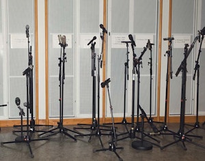10 mikrofoner i stativer står på rekke foran en grå vegg