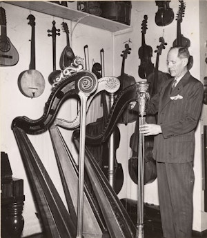 En eldre Trygve Lindeman betrakter harper i sin instrumentsamling. På veggen bak ham henger et utvalg strykeinstrumenter.