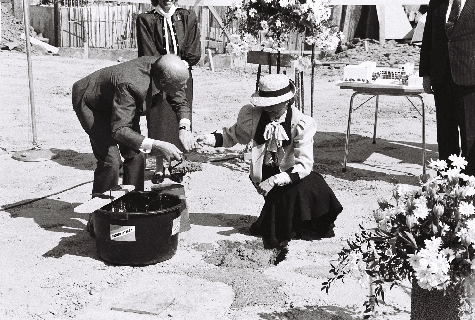 Kronprinsesse Sonja med hvit bredbremmet hatt sitter på huk ved siden av en bøtte med mørtel og skal legge ned grunnstein til første hus på Majorstua. Svart-hvitt.