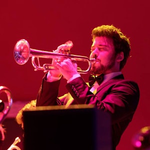 Alex Maestro står på en scene ikledd dress og spiller trompet